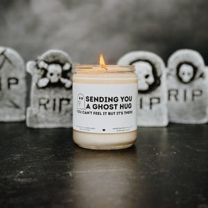 Puede incluir: Una vela encendida en un tarro de cristal con el texto "SENDING YOU A GHOST HUG" y una ilustración de un fantasma. La vela está frente a decoraciones de lápidas grises. Hecho a mano con cera de soja.