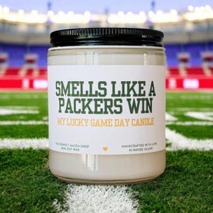 Puede incluir: Una vela en tarro de vidrio con tapa negra y etiqueta blanca. La etiqueta dice "Smells Like a Packers Win" en texto verde. La vela está sobre un campo verde con líneas blancas.