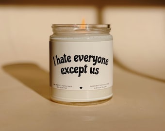 Pode incluir: Uma vela de cera de soja branca acesa num frasco de vidro. O rótulo diz "I hate everyone except us" em texto preto. A vela é feita à mão em Rhode Island.