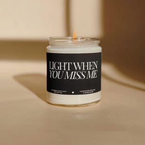 May include: A lit soy wax candle in a clear glass jar. The black label reads "LIGHT WHEN YOU MISS ME" in white text. The candle is handcrafted in Rhode Island.