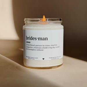 Puede incluir: Una vela de cera de soja encendida en un tarro de cristal transparente. La etiqueta dice "brides·man" con una definición. La vela está hecha a mano en Rhode Island. Un regalo temático de boda.