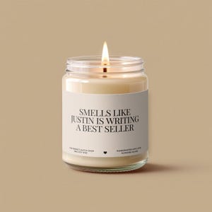 May include: A lit soy wax candle in a clear glass jar. The label reads "SMELLS LIKE JUSTIN IS WRITING A BEST SELLER." The candle is handcrafted in Rhode Island. The jar is on a neutral background.