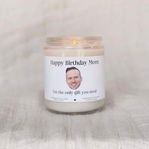 Op de afbeelding: Een glazen potkaars met een wit etiket. Het etiket zegt "Happy Birthday Mom" en "I'm the only gift you need", met een gezicht van een man. De kaars is lichtbeige en zit in een heldere glazen pot.
