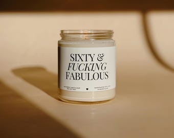 Può includere: Una candela in barattolo di vetro con un'etichetta bianca che recita "Sixty & Fucking Fabulous" in testo nero. La candela è riempita di cera cremosa e poggia su una superficie dai toni neutri.