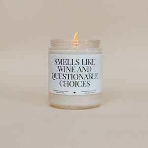 Puede incluir: Una vela de cera de soja encendida en un tarro de cristal transparente. La etiqueta dice "SMELLS LIKE WINE AND QUESTIONABLE CHOICES". La vela está hecha a mano en Rhode Island. Es un artículo de decoración del hogar.