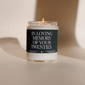 Op de afbeelding: Een brandende kaars in een glazen pot. Het zwarte etiket draagt de witte tekst: "IN LOVING MEMORY OF YOUR TWENTIES". Gemaakt van sojawas en handgemaakt in Rhode Island.
