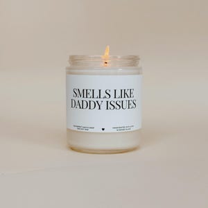 May include: A lit candle in a clear glass jar with a white label. The label reads "Smells Like Daddy Issues" in black text. The candle is from The Perfect Match Shop and is handcrafted with love in Rhode Island.