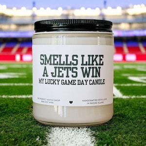 May include: A glass jar candle with a black lid and a white label. The label reads "SMELLS LIKE A JETS WIN" and "MY LUCKY GAME DAY CANDLE". The candle is on a green field with white yard lines.