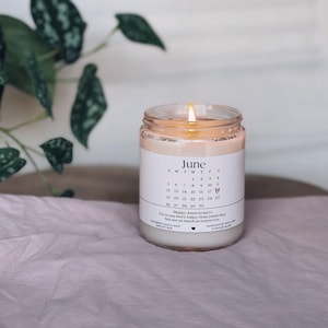 May include: A lit candle in a clear glass jar with a white label featuring a June calendar and the text "Happy Anniversary! I love you more today than yesterday, but not as much as tomorrow." The candle sits on a pink surface.
