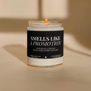 May include: A lit candle in a clear glass jar with a black label. The label says "Smells Like A Promotion" and "Congratulations! Your Hard Work Paid Off." The candle is made with soy wax and is on a neutral background.
