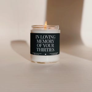 Puede incluir: Una vela encendida de cera de soja en un tarro de cristal transparente. La etiqueta negra dice "IN LOVING MEMORY OF YOUR THIRTIES". La vela es de The Perfect Match Shop y está hecha a mano en Rhode Island.