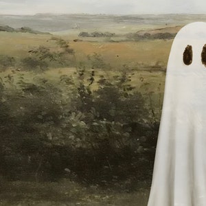 May include: A white ghost with two black eyes appears to be floating in front of a painting of a landscape with green bushes and a distant body of water.