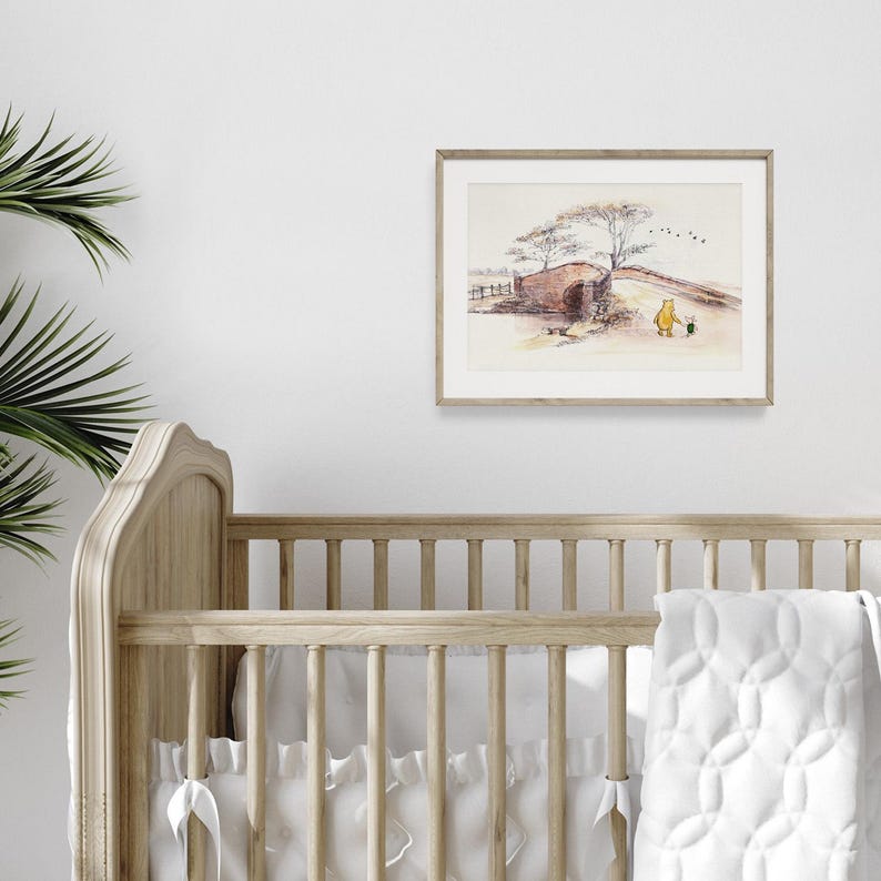 K&ouml;nnte beinhalten: Gerahmter Aquarelldruck mit einer Br&uuml;ckenszene mit einem gelben B&auml;ren und einer kleinen schwarzen Figur. Das Kunstwerk befindet sich in einem hellen Holzrahmen und h&auml;ngt &uuml;ber einem Holzbettchen mit einer wei&szlig;en Steppdecke. Der Druck hat eine weiche, ged&auml;mpfte Farbpalette.