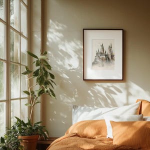 May include: A watercolour painting of Hogwarts castle, framed in a dark wood frame, hangs on a beige wall above a bed with orange bedding and white pillows.