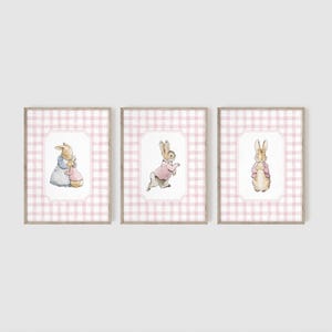 May include: Set of three framed watercolor prints featuring charming bunny illustrations. Each print showcases a bunny in a different pose, set against a pink and white gingham background. The frames are a light brown color, adding a touch of warmth to the artwork.