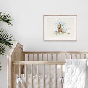 May include: A framed watercolor print featuring a cartoon character sitting on a tree stump. The artwork has a light blue sky background with black dots. The print is in a light wood frame and hangs above a wooden crib.