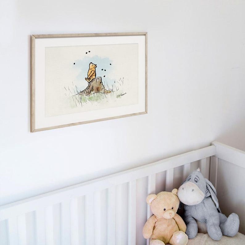 May include: Framed watercolor print featuring a yellow bear character sitting on a tree stump, with a light blue sky background and black accents. The artwork is in a light wood frame and is displayed above a white crib with stuffed animals.