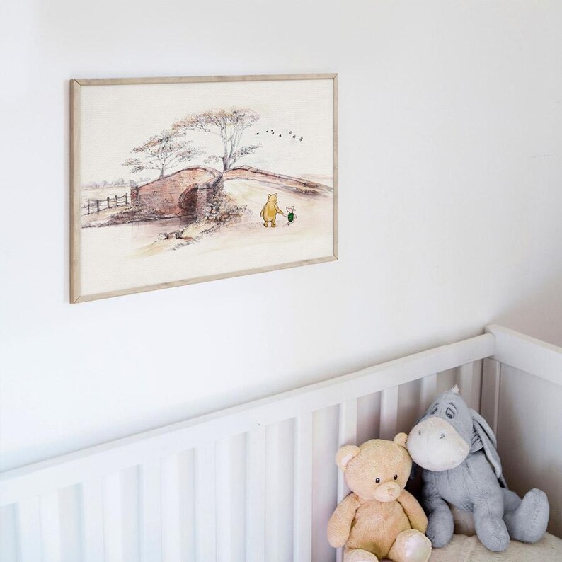 K&ouml;nnte beinhalten: Ein gerahmter Aquarelldruck zeigt eine klassische M&auml;rchenlandschaft mit einem B&auml;ren und einer kleinen Figur, die in der N&auml;he einer Steinbr&uuml;cke spazieren gehen. Das Kunstwerk befindet sich in einem hellen Holzrahmen und h&auml;ngt an einer wei&szlig;en Wand &uuml;ber einem Kinderbett.