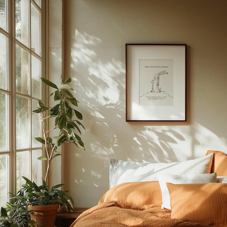 May include: A framed print of "The Velveteen Rabbit" with a black and white illustration of a rabbit. The print is in a dark brown frame, hanging on a beige wall. A bed with orange bedding and pillows is in the foreground, with a potted plant and window to the left.