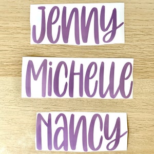 May include: Three stickers with names in a purple script font. The names are Jenny, Michelle, and Nancy. The stickers are on a white background and placed on a light brown wooden surface.