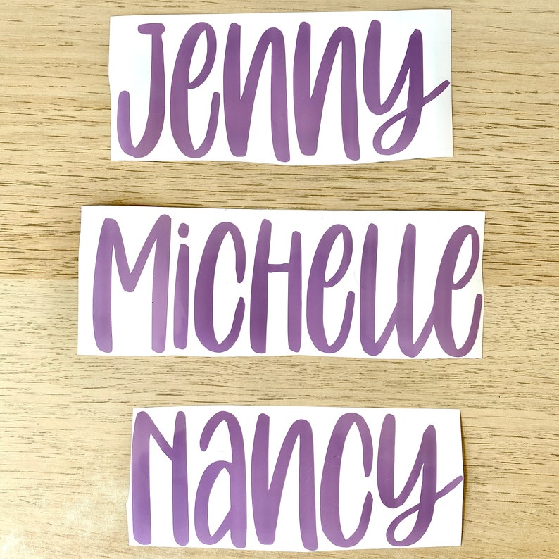 May include: Three stickers with names in a purple script font. The names are Jenny, Michelle, and Nancy. The stickers are on a white background and placed on a light brown wooden surface.