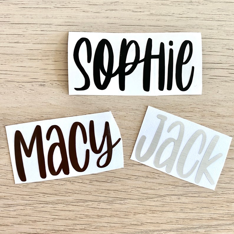 May include: Three name decals on a light wood surface. The top decal says "Sophie" in black cursive. Below it is "Macy" in brown cursive, and to the right is "Jack" in light grey cursive.