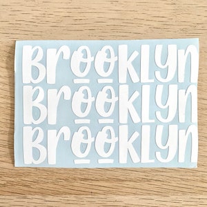 May include: White vinyl stickers with the name "Brooklyn" in a playful, rounded font. The stickers are arranged in three rows on a light blue backing, ideal for personalising items. The background is a light wood grain.