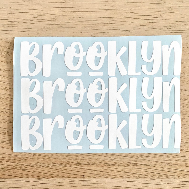 May include: White vinyl stickers with the name "Brooklyn" in a playful, rounded font. The stickers are arranged in three rows on a light blue backing, ideal for personalising items. The background is a light wood grain.