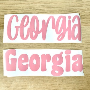 May include: Two pink vinyl decals with the name "Georgia" in different fonts. The top decal features a cursive font, while the bottom one uses a bubble letter style. Both are on a white background.