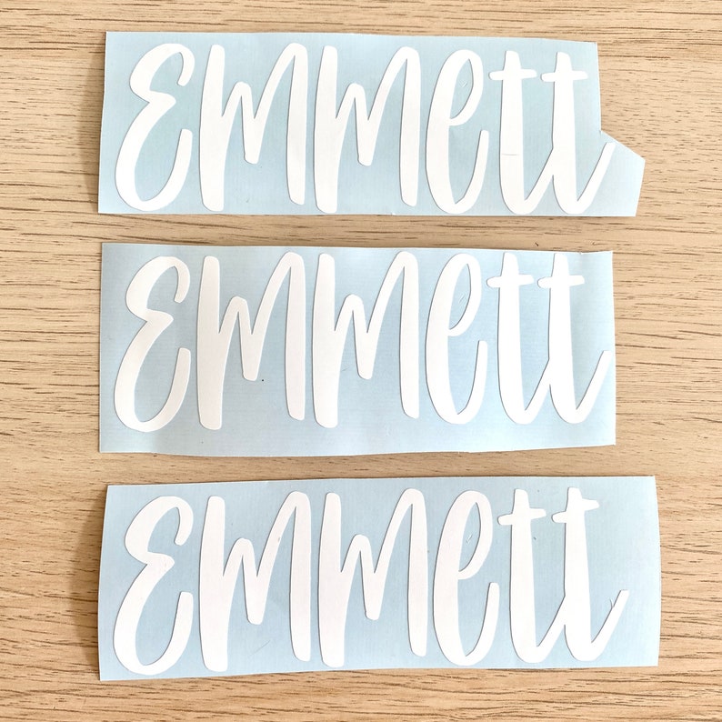 May include: Three white vinyl decals, each bearing the name "Emmett" in a cursive script. They are set against a light blue background. The background is a light wood grain.