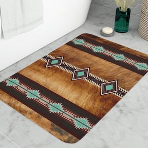 Puede incluir: Alfombra de baño rectangular con textura de piel sintética marrón y diseño de inspiración suroeste. El diseño presenta patrones geométricos turquesa, blancos y marrón oscuro. La alfombra está colocada frente a una bañera blanca.