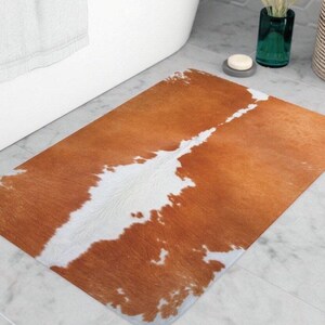 Puede incluir: Una alfombra de baño rectangular con un diseño de piel de vaca. La alfombra presenta un diseño marrón y blanco, con las secciones marrones dominando la superficie. Las secciones blancas tienen forma irregular, creando un aspecto naturalista. La alfombra está colocada frente a una bañera blanca.