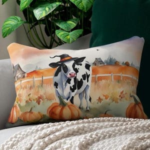 May include: Rectangular decorative pillow featuring a watercolor illustration of a cow wearing a witch hat in a fall landscape. The cow is black and white, surrounded by pumpkins and a fence. The background has a farmhouse and autumn colors.
