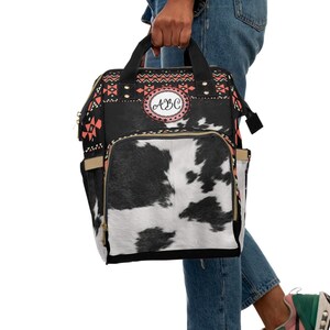 May include: A black and white cow print diaper bag with a red, black, and white geometric pattern on the top flap. The bag has a gold zipper and a gold handle. The bag has a circular patch with the letters "ABC" in pink.