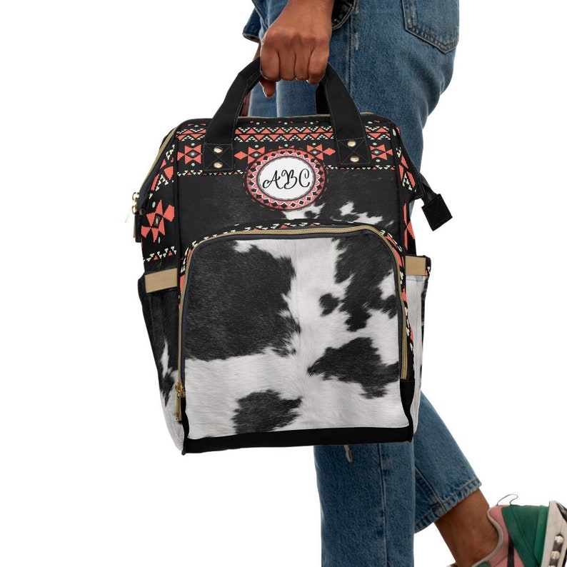 May include: A black and white cow print diaper bag with a red, black, and white geometric pattern on the top flap. The bag has a gold zipper and a gold handle. The bag has a circular patch with the letters "ABC" in pink.