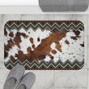 Puede incluir: Alfombra de baño rectangular con un estampado de piel de vaca en marrón, blanco y beige. La alfombra tiene un borde decorativo con un diseño en zigzag en marrón, blanco y turquesa. Un par de zapatillas grises en primer plano.