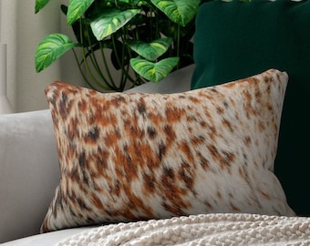 May include: Rectangular decorative pillow with a cowhide pattern in shades of brown, black, and white. The pillow is placed on a light gray sofa, with a green pillow and plant in the background. The pillow has a soft, textured appearance.