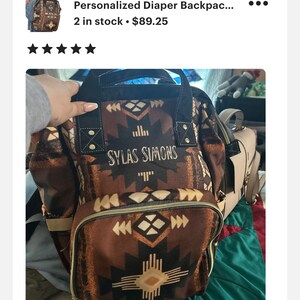 May include: A brown and black diaper backpack with a geometric pattern and the name "SYLAS SIMONS" embroidered on the front. The backpack has a large main compartment and several smaller pockets.