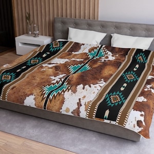 Op de afbeelding: Een bruin, wit en turquoise bedspreid met een koehuidprint. De bedspreid ligt op een bed met twee witte kussens.