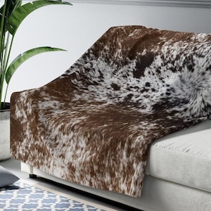 May include: A brown and white cowhide throw blanket with a soft, fuzzy texture. The blanket is draped over a white couch.