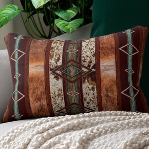 May include: Rectangular decorative pillow with a Southwestern design. The pillow features a brown, tan, and turquoise color scheme with geometric patterns and vertical stripes. The pillow is placed on a light-colored sofa with a cream-colored blanket.