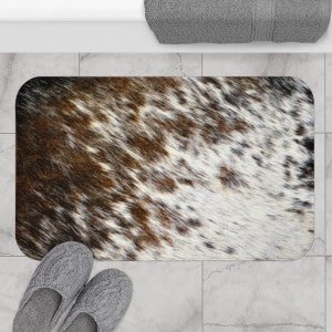 Puede incluir: Una alfombra de baño rectangular con estampado de piel de vaca marrón y blanca. La alfombra es suave y absorbente.