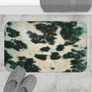 Puede incluir: Alfombra de baño rectangular con un estampado de piel de vaca en crema, negro y turquesa. La alfombra tiene esquinas redondeadas y está colocada sobre un suelo embaldosado. Zapatillas grises en la parte inferior izquierda.