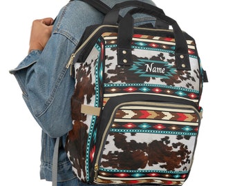 May include: A black and brown patterned diaper bag with a turquoise and red geometric design. The bag has a large front pocket and a zipper closure. The text "Name" is embroidered on the front of the bag.