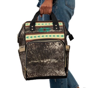 May include: A black and brown backpack with a cowhide pattern and a turquoise and brown geometric design. The backpack has a zippered pocket on the front and two adjustable straps.