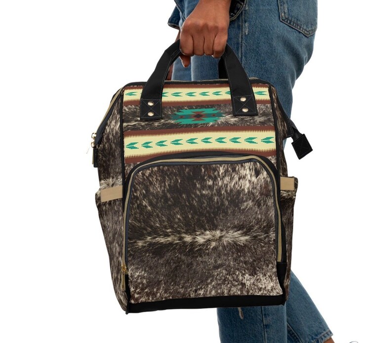 May include: A black and brown backpack with a cowhide pattern and a turquoise and brown geometric design. The backpack has a zippered pocket on the front and two adjustable straps.