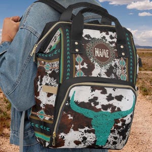 Personalized brown and white faux cowhide print with southwestern tribal design, western style diaper bag backpack. Custom monogrammed with your name or initials. Faux animal print backpack with a turquoise longhorn bull skull