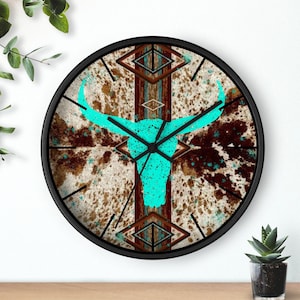 May include: Round wall clock with a black frame. The clock face features a turquoise bull skull design over a cowhide pattern with brown and turquoise accents. The clock hands are black.