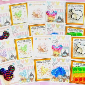 May include: A collection of assorted character-shaped pins and keychains displayed on individual cards. The items feature colorful designs including glittery mouse-shaped icons, lollipops, and white glove shapes, all set against a light pink background.