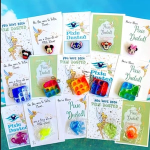May include: A collection of cards and small, colorful charms. The cards feature Tinkerbell illustrations and the text "Pixie Dusted!" and "You have been Pixie Dusted!". The charms include Mickey Mouse, Stitch, and other character designs.
